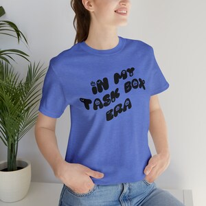 May include: Blue t-shirt with the text "IN MY TASK BOX ERA" in black, stylized lettering. The design incorporates pumpkin graphics. The shirt is a crew neck and has short sleeves.