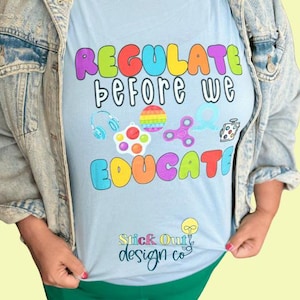 May include: Light blue t-shirt with the text "Regulate before we educate" in rainbow colors. The text is surrounded by colorful images of popular fidget toys. The bottom of the shirt has the text "Stick Out design co."
