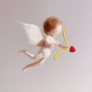 May include: A felt angel figurine with wings, holding a bow and arrow with a red heart. The angel is flying with its wings spread out.