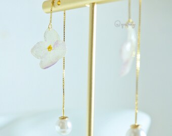 Op de afbeelding: Een paar delicate oorbellen met een witte bloem met paarse accenten en een parel hanger. De oorbellen worden getoond op een gouden standaard.