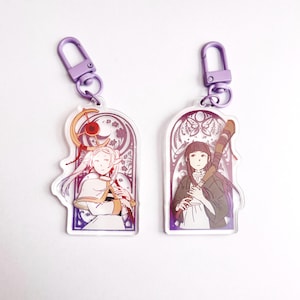 May include: Two clear acrylic keychains with purple lobster clasps. The left charm features a light-haired character with a staff in a decorative archway. The right charm depicts a dark-haired character holding a wooden staff within a similar frame.