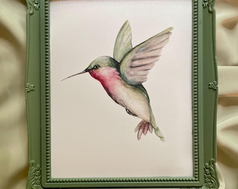 May include: A framed watercolor painting of a hummingbird in flight. The bird has a red throat, green back, and gray wings. The frame is a muted green with ornate detailing.