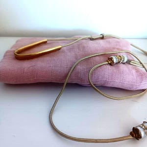 May include: A gold-toned necklace with a U-shaped pendant and a delicate chain. The necklace features white and gold accents, resting on a pink fabric. The jewelry is displayed on a white surface.