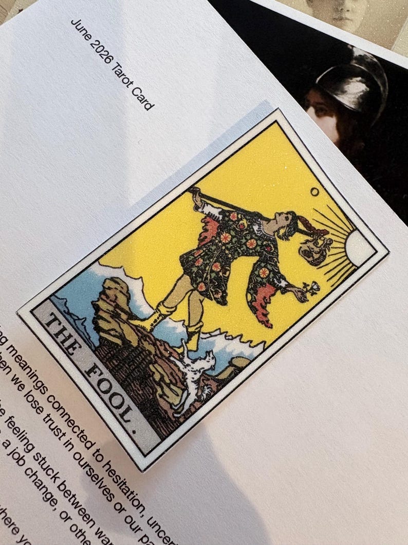 Pu&ograve; includere: Una carta dei tarocchi rettangolare intitolata THE FOOL che raffigura una figura in tunica floreale in piedi su una scogliera con un piccolo cane bianco, su uno sfondo giallo brillante con un sole, stampata su un foglio di carta bianco con il testo June 2026 Tarot Card.