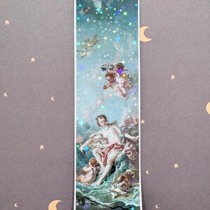 以下が含まれることがあります： 青い空と海を背景に、中央の人物と周囲の天使が描かれたクラシックな絵画の長方形のしおり。表面にはホログラムの星模様が施されており、光を反射して様々な色に輝くデザインとなっている。