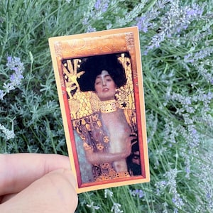 Peut inclure: Un autocollant rectangulaire représentant la peinture Judith et Holopherne de Gustav Klimt. L'œuvre montre une femme aux cheveux sombres, portant un collier ras de cou doré et des vêtements à motifs dorés, avec le texte JUDITH AND HOLOFERNES en haut.