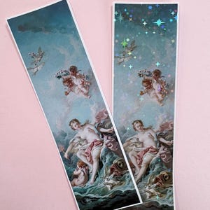 以下が含まれることがあります： 雲と海泡の中に横たわる人物、天使、鳥を描いた古典的な絵画がプリントされた2枚の長方形のブックマーク。片方のブックマークには、光を反射するホログラフィックの星のオーバーレイ加工が施されています。