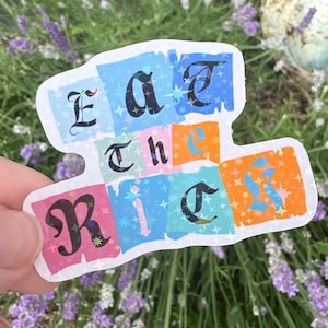 May include: A die-cut sticker with a holographic star pattern featuring the text Eat the Rich. The design consists of colourful rectangular blocks in blue, orange, and pink, with black serif lettering layered over the top of the vibrant background.