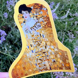 May include: A die-cut sticker featuring a reproduction of Gustav Klimt's Portrait of Adele Bloch-Bauer I. The artwork displays a woman with dark hair in a gold-toned gown adorned with intricate geometric patterns, eye motifs, and metallic gold leaf.
