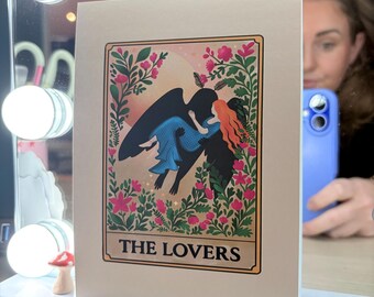 May include: A tarot card featuring an illustration of two figures embracing, surrounded by floral elements. The card is labeled "THE LOVERS" in a rectangular frame. The figures are a person with red hair and a person with black wings and a blue dress. The card is displayed on a white surface.