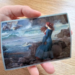 May include: A rectangular print of a painting showing a woman in a blue dress with red hair, on a rocky shore, looking at a stormy sea with a shipwreck. The artwork has a classical style with dramatic lighting and colour.