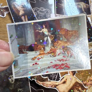 Puede incluir: Una pegatina rectangular con una pintura cl&aacute;sica de una figura con vestido blanco rodeada de leones y tigres en escalones de piedra, superpuesta con un patr&oacute;n de estrellas hologr&aacute;ficas. Incluye p&eacute;talos de flores rojas esparcidos.