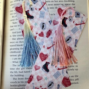Puede incluir: Dos marcapáginas decorativos con borlas descansan sobre un libro abierto. Los marcapáginas presentan un patrón de cuadros rosas y blancos con corazones, patines de hielo y palos de hockey. Una borla es azul y amarilla, la otra es rosa.