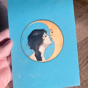 Op de afbeelding: Een turquoise wenskaart met een vintage illustratie van een vrouw die een halve maan kust. De vrouw heeft donker haar en staat tegen een gouden maan. De kaart heeft een gespikkelde textuur.