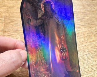 Puede incluir: Una pegatina holográfica alta y arqueada que presenta una reproducción del cuadro La luz del mundo. La imagen muestra a una figura con túnica y halo en un bosque oscuro, sosteniendo una linterna encendida y llamando a una puerta.