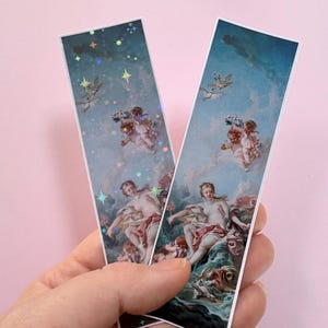 以下が含まれることがあります： 雲と青空の中に横たわる人物と天使が描かれた古典的な絵画の長方形のしおり2枚。左側のしおりにはホログラムの星模様が重ねられており、右側のしおりには星模様のない同じ絵柄が描かれています。