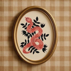 May include: Oval gold framed artwork featuring a pink snake illustration. The snake is decorated with red leaf patterns and surrounded by black leaves and flowers against a cream background. The artwork is displayed on a tan and white checkered surface.