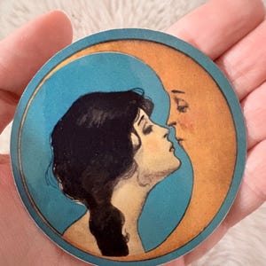 Op de afbeelding: Een ronde sticker met een vintage illustratie van een vrouw die de maan kust. De vrouw heeft donker haar en is in profiel, kussend een halve maan met een lichtoranje tint. De achtergrond is helderblauw.