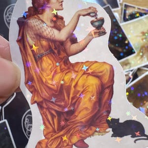Puede incluir: Una pegatina troquelada con una ilustraci&oacute;n cl&aacute;sica de una persona de cabello rojizo con una t&uacute;nica dorada y naranja, sosteniendo una botella peque&ntilde;a y un c&aacute;liz, con un gato negro a sus pies, todo cubierto con un patr&oacute;n de estrellas hologr&aacute;ficas.
