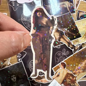 Puede incluir: Una colecci&oacute;n de pegatinas con reproducciones de arte cl&aacute;sico y un acabado hologr&aacute;fico de estrellas. La pegatina central muestra una figura con capa roja. Otras pegatinas muestran varias figuras, un tigre y una criatura alada con un efecto brillante.