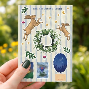 Peut inclure: Une planche d'autocollants au design fantaisiste avec deux lièvres bruns sautant, une couronne florale, des étoiles et un croissant de lune. Le texte "THE RETURNING LIGHT" est en haut. D'autres autocollants incluent un paysage, une fleur et une scène de nuit.