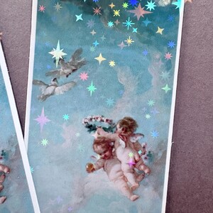 以下が含まれることがあります： 水色の空を背景にホログラムの星が散りばめられた長方形のしおり。花輪を持つ2人の翼のある天使、飛んでいる2羽の白い鳥、そして下部には顔の一部が描かれています。