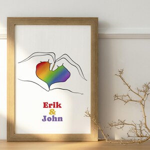 Puede incluir: Una impresión enmarcada que presenta una forma de corazón formada por manos, con un corazón de colores del arcoíris en el centro. Los nombres "Erik & John" están impresos debajo en rojo y morado. El marco es marrón claro.
