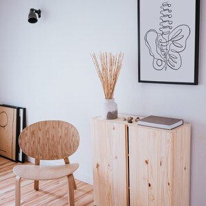 May include: A wooden cabinet with two doors, a light wood chair, a vase with dried bamboo stalks, and a framed black and white line drawing of a butterfly. The cabinet is on a wood floor.