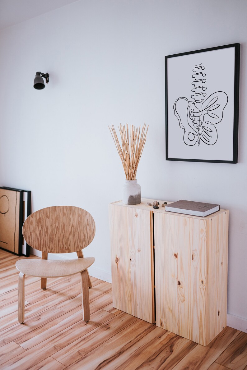 May include: A wooden cabinet with two doors, a light wood chair, a vase with dried bamboo stalks, and a framed black and white line drawing of a butterfly. The cabinet is on a wood floor.