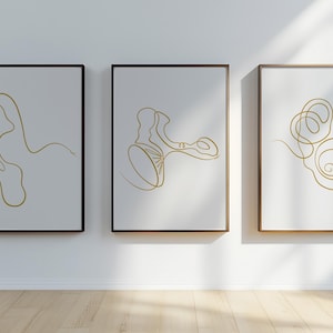 Può includere: Tre stampe di arte minimalista incorniciate con linee dorate su uno sfondo bianco. Le stampe sono in cornici nere e sono appese a un muro bianco.