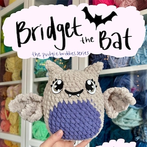 May include: A crocheted bat plush toy named "Bridget the Bat" from the "Pudge Buddies" series. The toy is gray with purple wings and has large, sparkly eyes.