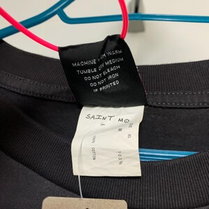 May include: Black t-shirt with a white tag that says "SAINT MO" and "100% COTTON". The tag also has a size label that says "XL". There is a smaller tag that says "SAINT M" and "NO. SM-S22-0000-074".