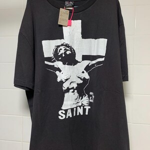 May include: Black t-shirt with a white graphic of a person on a cross. The graphic includes the word "SAINT" below the figure.