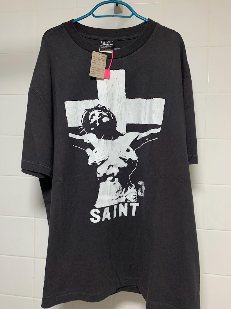 May include: Black t-shirt with a white graphic of a person on a cross. The graphic includes the word "SAINT" below the figure.