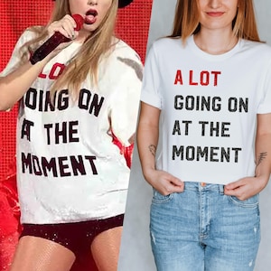 以下が含まれることがあります： 黒と赤で「A LOT GOING ON AT THE MOMENT」とプリントされた白いTシャツ