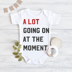 May include: White baby onesie with the text "A LOT GOING ON AT THE MOMENT" in black and red lettering.
