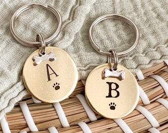 May include: Two gold-colored dog tags with key rings. The tags are engraved with the letters "A" and "B" and a paw print. Each tag has a small silver bone charm.