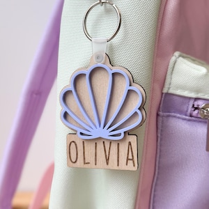 May include: A wooden keychain with a seashell design and the name "OLIVIA" engraved on it. The seashell is light brown with a light purple outline. The keychain is attached to a backpack with a silver clasp.