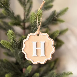 May include: A wooden Christmas decoration featuring the letter "H" in white. The ornament has a scalloped edge with a white border, suspended by twine. It hangs against a backdrop of a green Christmas tree.