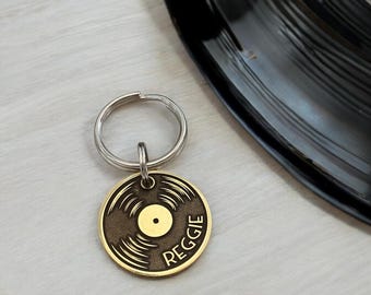 May include: A gold-colored, circular pet tag with a vinyl record design. The tag has the name "Reggie" engraved on it. A silver-colored key ring is attached to the tag. A black vinyl record is in the background.