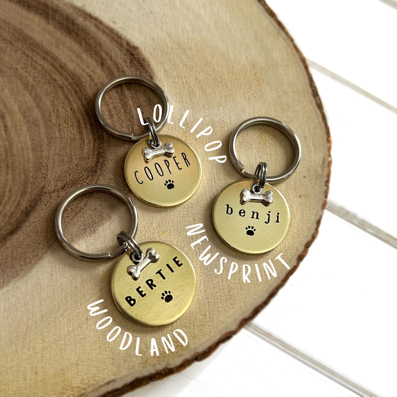 May include: Three gold dog tag keychains with engraved names "Cooper", "Bertie", and "Benji". Each tag features a small silver bone charm and a paw print. The tags are arranged on a wooden surface.