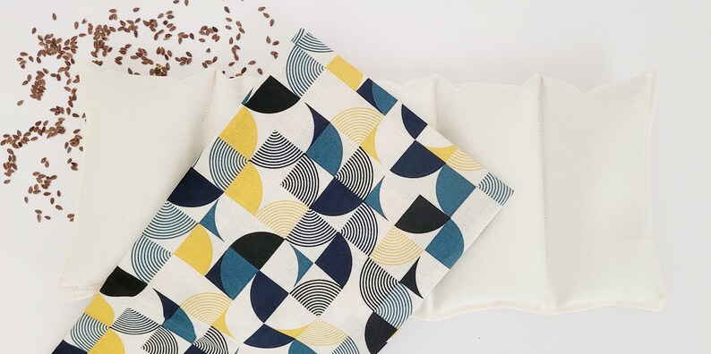 May include: Two white linen flax seed heating pads, one with a geometric pattern of blue, yellow, and black circles, and one with a plain white surface.