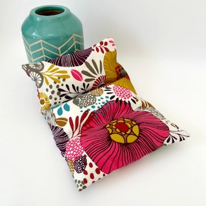 Peut inclure: Un coussin en tissu blanc avec un motif floral coloré dans des tons de rose, jaune, violet et turquoise. Le coussin est plié en deux et a une surface légèrement texturée.