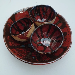 Pode incluir: Quatro tigelas pequenas de cerâmica com um padrão de esmalte vermelho, preto e azul. As tigelas estão aninhadas dentro de uma tigela maior com o mesmo padrão de esmalte.