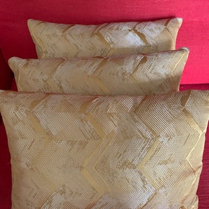 May include: Three gold throw cushions with a chevron pattern. The cushions are stacked on a red sofa.
