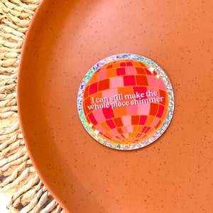 May include: A round sticker with a glittery border and a pink and orange disco ball design. The text "I can still make the whole place shimmer" is printed on the sticker.