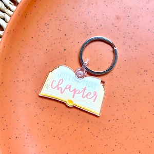 May include: A clear acrylic keychain shaped like an open book with the text "Just one more chapter" in pink and white lettering. The keychain is attached to a silver metal ring.