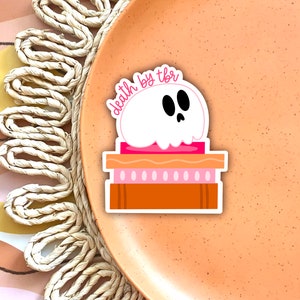 May include: A white ghost with black eyes sits on top of a pink and brown layered cake. The text "death by tbr" is written in pink above the ghost.
