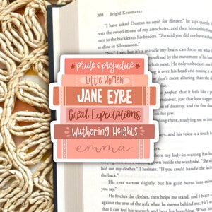 May include: A sticker featuring a stack of books with titles like "Pride & Prejudice," "Jane Eyre," and "Great Expectations." The books are in shades of pink and peach, with the titles in a mix of fonts. The sticker is placed on an open book.