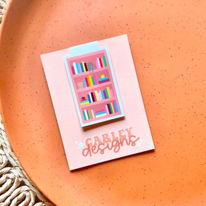 May include: A pink and white bookmark with a colorful illustration of a bookshelf filled with books. The bookmark has the text "CARLEY designs" on the bottom.